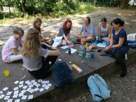 Eine Gruppe junger Personen sitzt im Kreis auf dem Boden. Um sie herum liegen verschiedene Zettel, Stifte und Snacks.