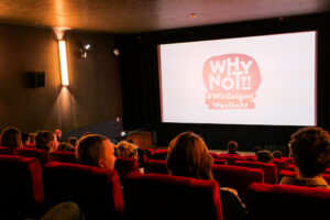 Ein Kinosaal in dem Menschen sitzen, auf der Leinwand ist ein Logo mit der Aufschrift "WhyNOT?! - #wirzeigenwasgeht"