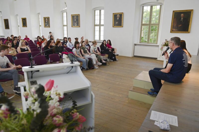 Zwei Personen sitzen am Rand einer Bühne vor einem Publikum aus jungen Menschen.