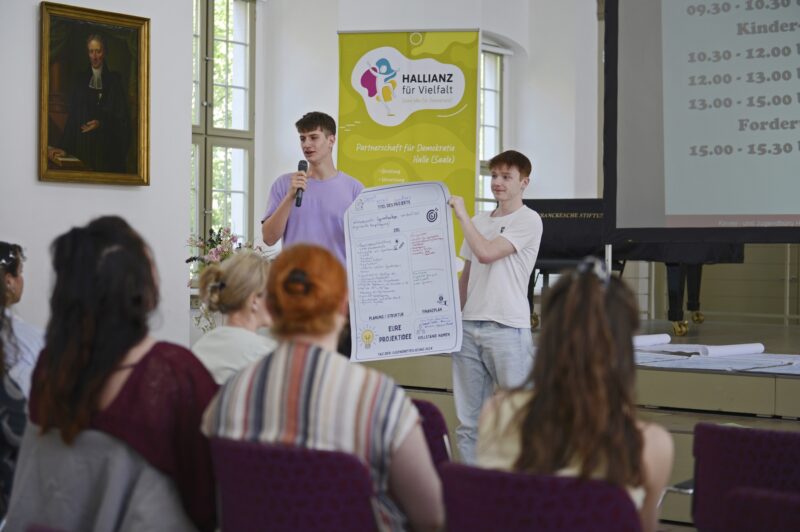 Zwie junge Personen stehen vor einem Publikum, die eine hält ein Plakat, die andere spricht in ein Mikrofon.